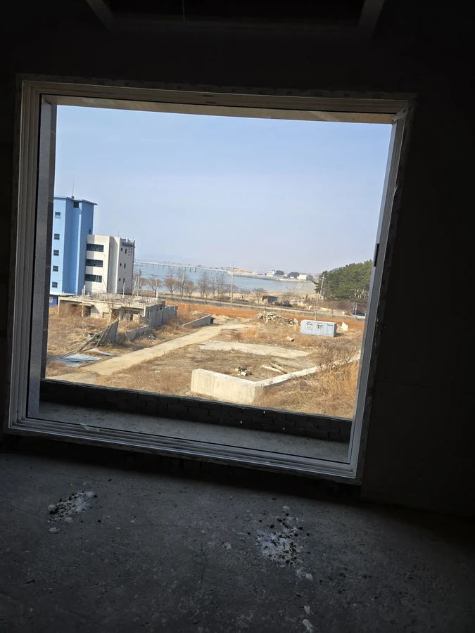 어두운 실내에서 큰 창틀 너머로 바깥 부지와 건물이 보이는 모습